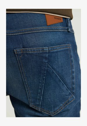 Jeans dżinsowe w ciemnym niebieskim kolorze z pomarańczowymi stębnowaniami. Posiadają charakterystyczny design kieszeni oraz skórzaną naszywkę z napisem "CHASIN'" na talii.