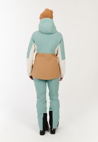 Sneeuwjack en -broek in teal, beige en crème. Het jack heeft een capuchon en elastische manchetten; de broek heeft zwarte accenten en is aansluitend bij de enkels.