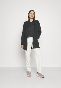 Schwarze Fleecejacke mit hohem Kragen, Reißverschluss und lässiger Passform. Kombiniert mit cremfarbenen Hosen und helllila Sportschuhen.
