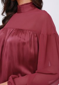 Burgunderfarbene Bluse mit hohem Kragen, aus leichtem, seidig weichendem Stoff. Verfügt über geraffte Details und lange, fließende Ärmel.