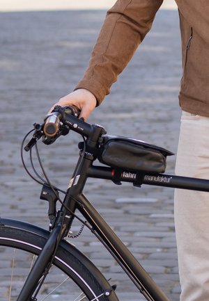 Eine Person in einer braunen Jacke hält den Lenker eines schwarzen Rennrades, an dessen Rahmen eine kleine schwarze Tasche angebracht ist.