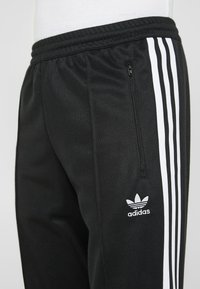 Svarta träningsbyxor i glatt tyg med ribbad midja, med vita sidostripes och en ficka med dragkedja. Adidas-logotypen synlig.