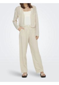 Cardigan de punto color crema con tejido abierto, combinado con pantalones beige de pierna ancha. Sandalias planas blancas completan el conjunto.