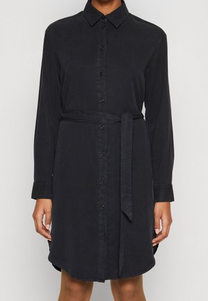 Vrouw draagt een zwarte jurk met lange mouwen, een kraag, knoopsluiting aan de voorkant en een tailleband, staand tegen een neutrale achtergrond.