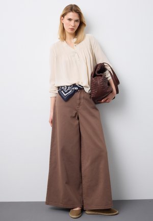 Vrouw draagt een crèmekleurig blouse, brede bruine broek, marineblauwe bandana in de taille, houdt een grote bruine geweven tas vast, staat tegen een witte muur.