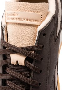 Καφέ δερμάτινο αθλητικό παπούτσι με υφή στον καφέ γλωσσάριο, που διαθέτει ετικέτα λογότυπου, υφασμάτινα κορδόνια και διακριτό σχέδιο γραναζιού στο υλικό.