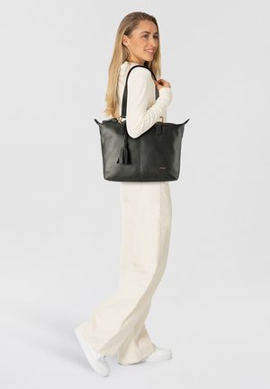 Black leather tote bag with a textured finish, features gold hardware, tassel detail, and dual handles. Model wears light-colored clothing.