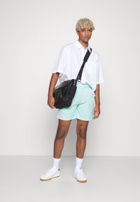 Sort messenger taske, hvid kortærmet skjorte, lysegrønne shorts og hvide sneakers med blå accenter. Glatte stoffer, afslappet pasform.