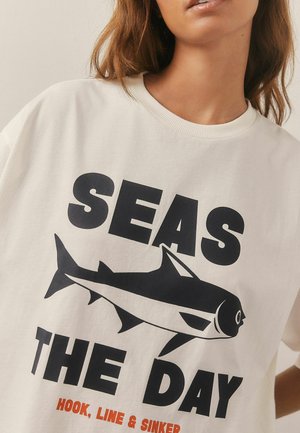 Valge puuvillane T-särk, millel on must graafiline tekst "SEAS THE DAY" ja hai illustreerimine, koos oranžide aktsentidega teksti all.