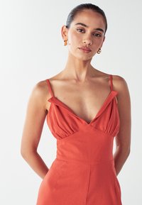 Žena s tmavými vlasmi stiahnutými dozadu, oblečená v zlatých kruhových náušniciach a bezrukávových červených šatách s tenkými ramienkami pred bielym pozadím.