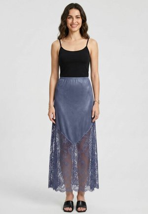 Femme aux longs cheveux ondulés porte un haut noir sans manches et une jupe maxi en dentelle bleue avec un panneau triangulaire solide, debout et souriante.