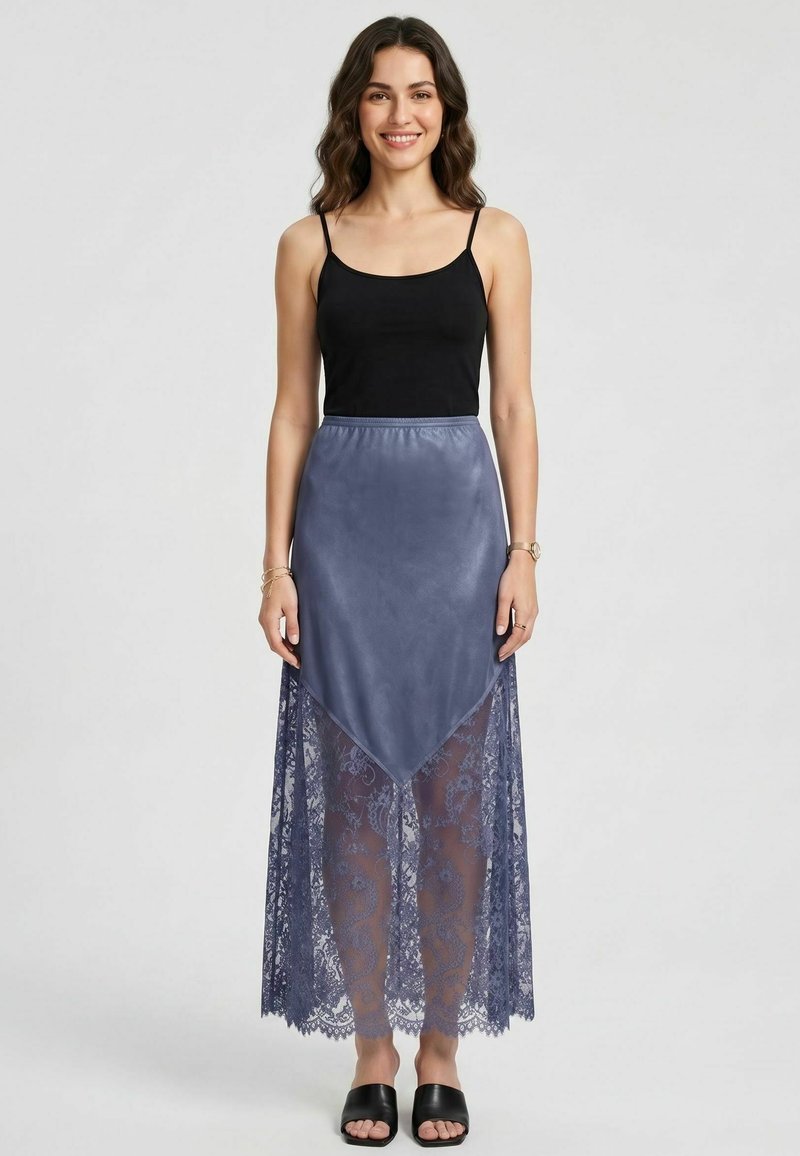 Femme aux longs cheveux ondulés porte un haut noir sans manches et une jupe maxi en dentelle bleue avec un panneau triangulaire solide, debout et souriante.