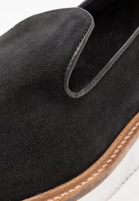 Chaussure noire en daim à enfiler avec un dessus arrondi, un détail découpé et une semelle en cuir naturel contrasté avec une bordure en caoutchouc blanc.