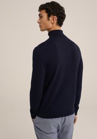 Tumši zila džemperis ar augstu apkakli, adīti raksti un garām piedurknēm. Modelis parādīts no aizmugures, ar gaiši pelēkiem adītiem biksēm.