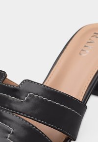 Gros plan sur une sandale à talon bloc en cuir noir avec couture blanche et semelle beige.