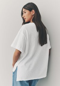 White oversized t-shirt with rolled sleeves, soft texture, side slits, paired with light blue denim jeans, viewed from the back.