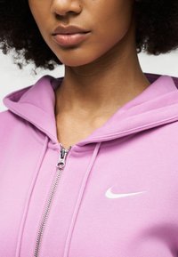 Νεαρός φοράει ανοιχτό μοβ φούτερ Nike με φερμουάρ και λευκό λογότυπο Swoosh, εμφανίζονται λεπτομέρειες του λαιμού και του κάτω μέρους του προσώπου.