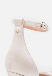 Beige satin wedge sandal with an ankle strap, featuring a gold-tone heart accent and stitched detailing on the heel.