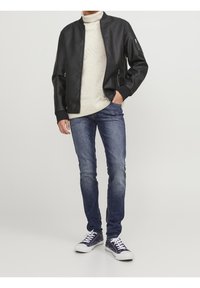 Giacca bomber in pelle nera, dolcevita beige, jeans blu e sneakers blu navy. La giacca presenta una chiusura con zip e polsini a coste.