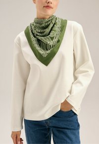 Biała bluzka z długim rękawem w połączeniu z zielonym jedwabnym szalikiem w wzór paisley. Szal ma kwadratowy kształt z kontrastującą ramką.