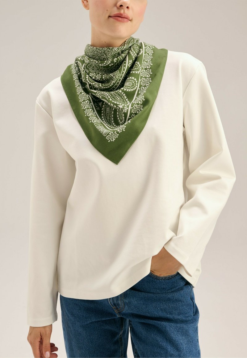 Biała bluzka z długim rękawem w połączeniu z zielonym jedwabnym szalikiem w wzór paisley. Szal ma kwadratowy kształt z kontrastującą ramką.