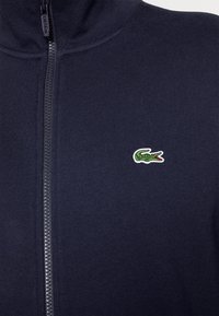 Veste zipée marine en tissu tricoté, avec un col haut et un petit logo crocodile brodé sur la poitrine gauche.