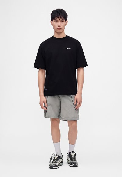 Νέος άνδρας που φοράει μαύρο υπερμεγέθες t-shirt, γκρι σορτς, λευκές κάλτσες μέχρι τον αστράγαλο και ασπρόμαυρα αθλητικά παπούτσια, στέκεται μπροστά σε ένα απλό φόντο.