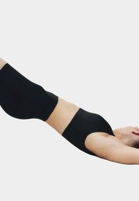 Žena leži na leđima s podignutim nogama, odjevena u crni sportski grudnjak bez šavova i kratke hlače na jednobojnoj svijetloj pozadini.