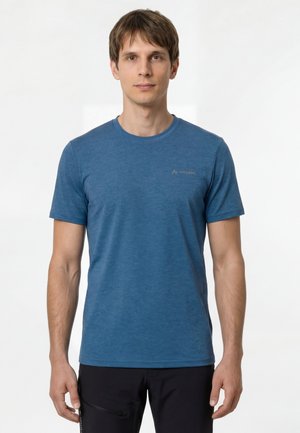 Junger Mann mit kurzen braunen Haaren, der ein schlichtes blaues kurzärmliges T-Shirt und schwarze Hosen trägt und vor einem weißen Hintergrund steht.