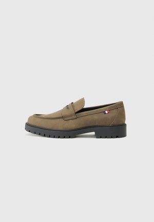 Mocasines de ante marrón con punta redondeada, suela de goma negra y costuras decorativas. Presenta un pequeño acento de bandera roja, blanca y azul.