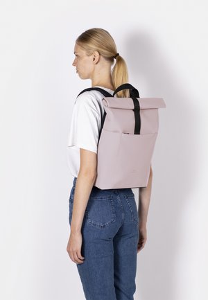 Woman with blonde hair in a ponytail wearing a white t-shirt and blue jeans carries a pink roll-top backpack with black straps on her back.