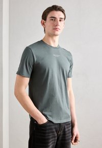 Grå crew-neck t-skjorte i mykt bomullsstoff, tettsittende design, med en liten logo foran. Kombinert med mørke jeans med et subtilt stripete mønster.