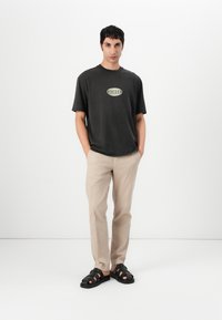 Mørk grå t-skjorte med grønt logo, beige bukser og svarte sandaler. T-skjorten har en avslappet passform og korte ermer.