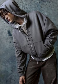 Felpa grigia con cerniera e cappuccio, dalla texture liscia, con tasche frontali e dettagli a costine all'orlo, indossata sopra una camicia bianca e dei pantaloni di velluto a coste marroni.