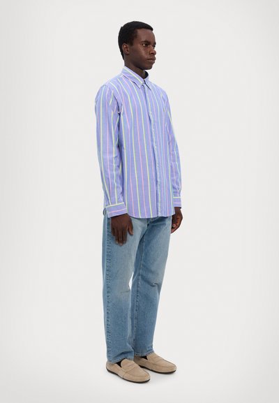 Chemise boutons jusqu'en violet clair avec des rayures verticales jaunes et vertes. Associée à un jean bleu et des chaussures décontractées beiges. Détail horizontal sur les poignets.