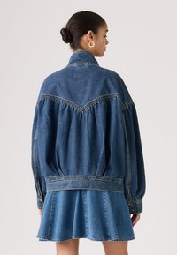 Γυναίκα με μαλλιά πιασμένα κότσο, που φοράει υπερμεγέθη μπλε τζιν μπουφάν και matching φούστα, εμφανιζόμενη από πίσω σε απλό φόντο.