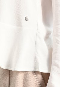 Top bianco strutturato con orlo corto, caratterizzato da un logo rotondo argentato sul lato sinistro. Tessuto morbido abbinato a un indumento beige sottostante.