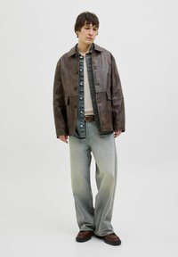 Brown leather jacket with a collared design, over a blue denim shirt, paired with light gray jeans and dark brown shoes. Minimalistic style.