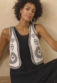 Gilet sans manches en crème avec broderie noire, présentant des motifs de diamants et floraux. Porté sur une robe en dentelle noire, mettant en valeur une texture complexe.