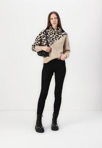 Un pull beige et noir à motifs avec un col bénitier, associé à un jean skinny noir et des bottes noires à lacets épaisses. Le mannequin tient un petit sac noir.