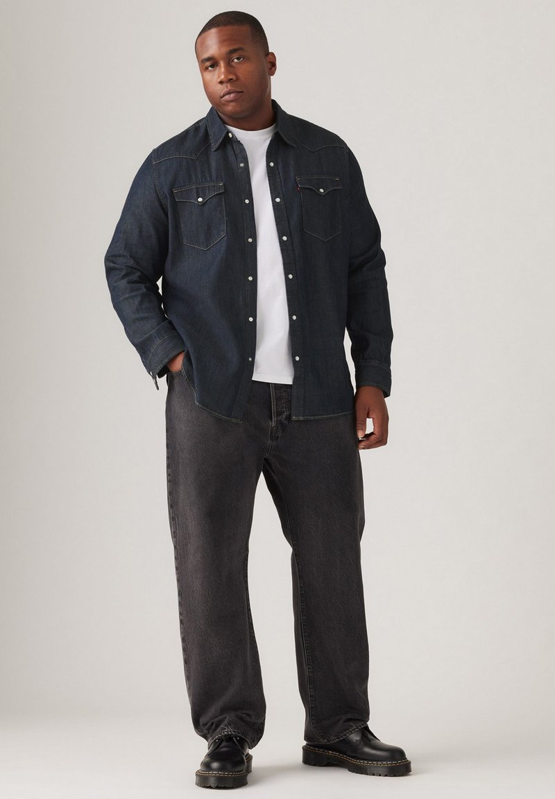 Chemise en denim foncé avec des poches poitrine, portée sur un t-shirt blanc, associée à un jean noir ample et des bottes noires.