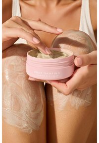 Barattolo rosa etichettato "Crema Fango Drenante Modellante per il Corpo" tenuto in una mano, con un prodotto cremoso e verdastro all'interno. Crema applicata su pelle abbronzata.