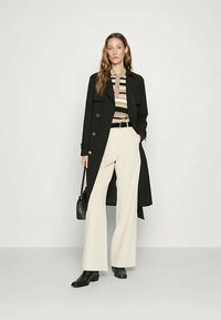 Mujer vestida con un abrigo negro, camiseta de rayas beige y negras, pantalones blancos de pierna ancha, botas negras, y sosteniendo un bolso negro, todo esto frente a un fondo liso.