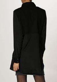 Camicia lunga nera realizzata in tessuto morbido, con colletto, polsini con bottoni e orlo leggermente curvo, che presenta un design minimalista.
