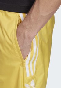 Gula sportshorts med vita ränder, tillverkade av lättviktig tyg. Har en sidficka och en broderad logotyp nära nederkanten.