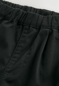 Pantalons noirs avec une taille élastique froncée, présentant un tissu lisse et plat, des détails de couture subtils et des poches inclinées sur les côtés.