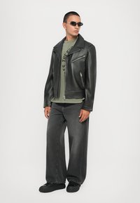 Μαύρο δερμάτινο μπουφάν μηχανής, πράσινη μπλούζα με σχέδιο, χαλαρό σκούρο τζιν και μαύρα παπούτσια. Τα αξεσουάρ περιλαμβάνουν μαύρα γυαλιά ηλίου.