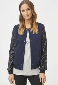 Chaqueta de-varsity azul marino con mangas de cuero negro, botones de presión, puños acanalados y bolsillos laterales, llevada sobre una camiseta gris.