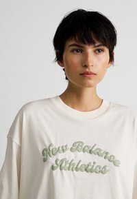 Person med kort mørkt hår som har på seg en hvit sweatshirt med teksten "New Balance Athletics" i hevede grønne bokstaver på forsiden.