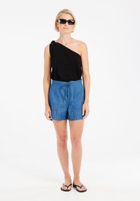Top negro de un hombro, pantalones cortos tipo jogging en azul vaquero con cordón y bolsillos laterales, y chanclas negras. Tela suave y ajuste relajado.